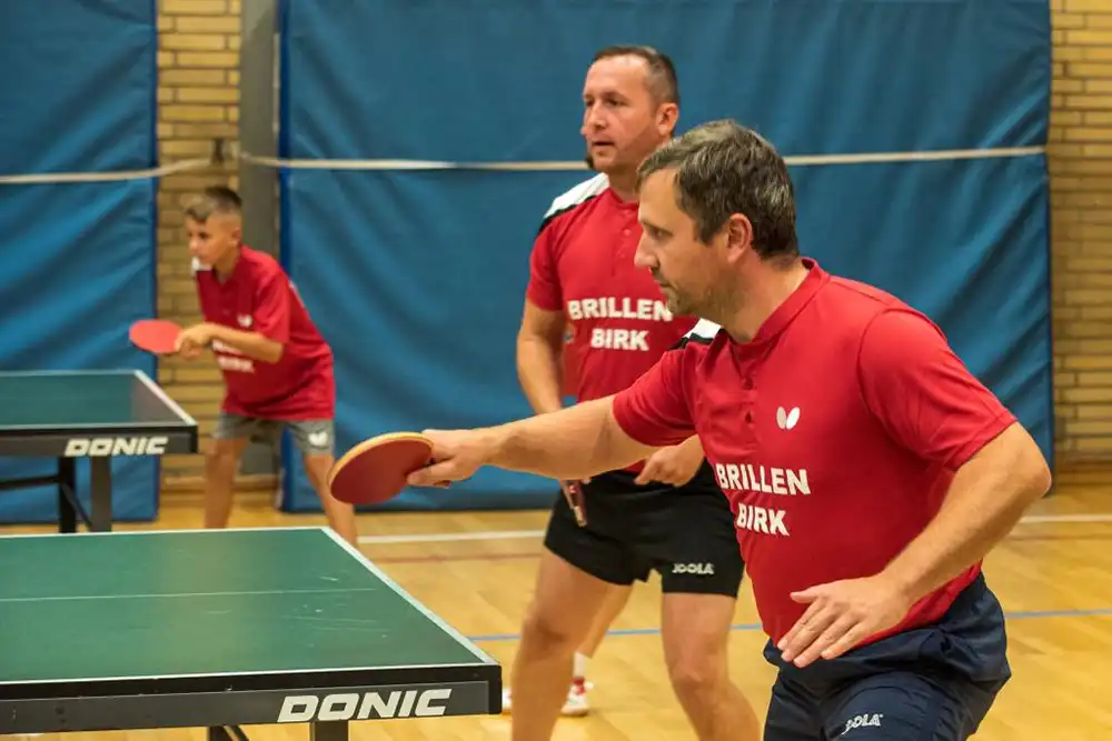 Drei Personen spielen Tischtennis in einem Sportraum. Zwei Männer stehen an einem Tisch, während ein Junge im Hintergrund ebenfalls spielt. Alle tragen rote Shirts mit der Aufschrift "BRILLEN BIRK".