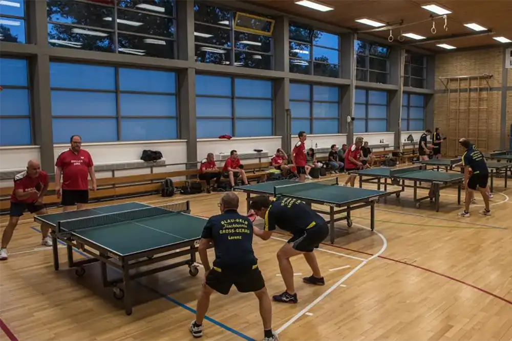 Tischtennistraining in der Halle mit Spielern des VfR Simmern; zwei Teams spielen an nebeneinanderstehenden Tischen, Zuschauer in roten Trikots sitzen im Hintergrund.