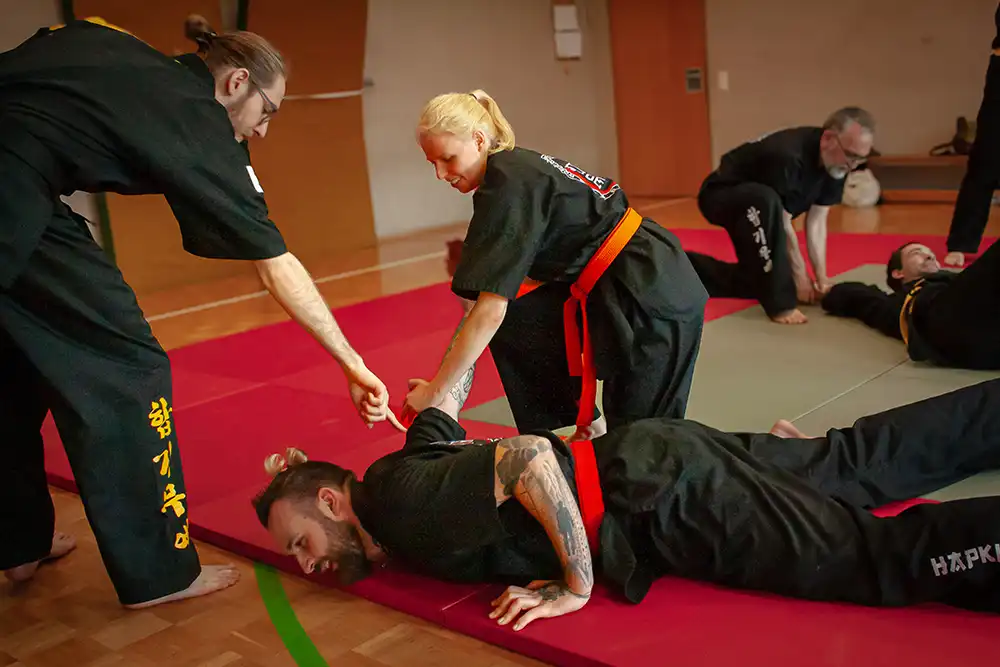 Ein Hapkido-Training mit mehreren Praktizierenden in schwarzer Kleidung, die verschiedene Kicks ausführen, während sie auf einer Trainingsmatte stehen.