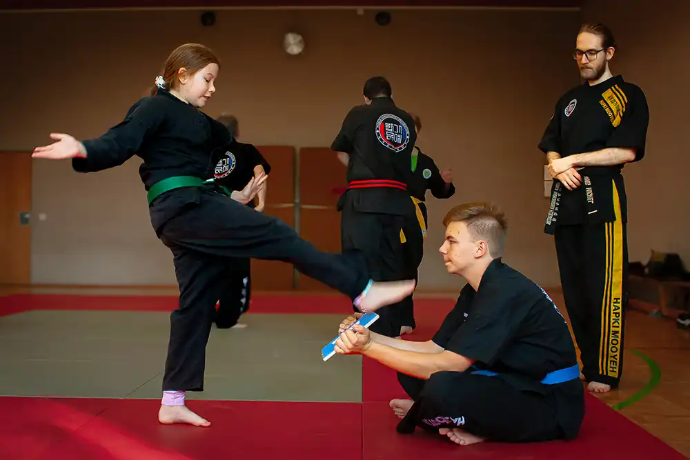Ein Kind mit grünem Gürtel führt einen Kick im Hapkido-Training aus, während ein anderer Schüler mit blauem Gürtel auf dem Boden sitzt und ein Trainingsgerät hält. Im Hintergrund sind weitere Teilnehmer, die ebenfalls in Hapkido-Uniformen trainieren.