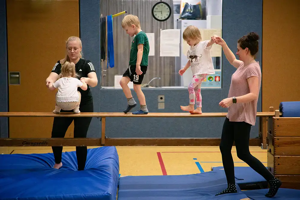 Zwei Kinder balancieren auf einem Schwebebalken, während eine Trainerin sie unterstützt. Eine weitere Erwachsene steht daneben und hilft einem Kind, das auf dem Balken geht.