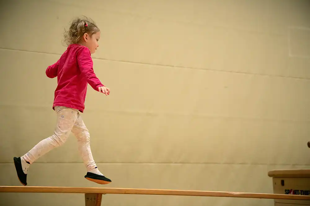 Ein kleines Mädchen in einem roten Shirt und hellen Leggings balanciert auf einem Turnbalken beim Eltern-Kind-Turnen des VfR Simmern.
