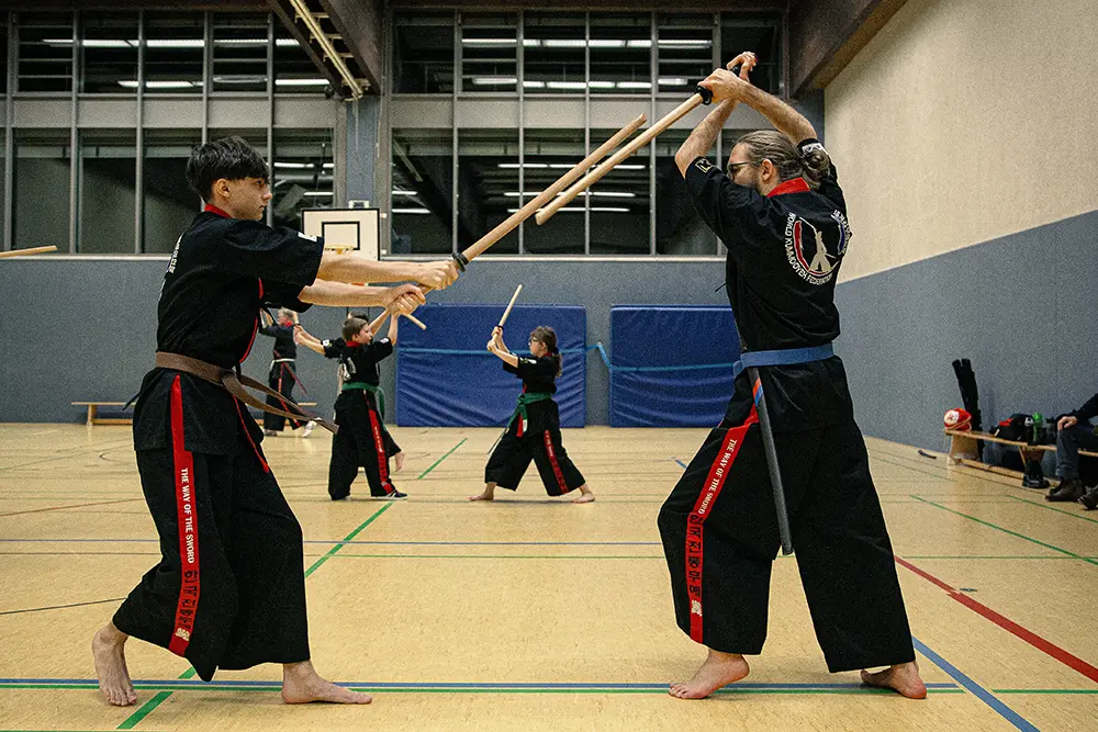 Zwei Kämpfer im Kummooyeh, einer koreanischen Kampfkunst, führen Schwertkampf-Übungen in einer Sporthalle durch. Der vordere Kämpfer, mit schwarzem Anzug und braunem Gürtel, greift an, während der andere Kämpfer mit einem blauen Gürtel verteidigt.