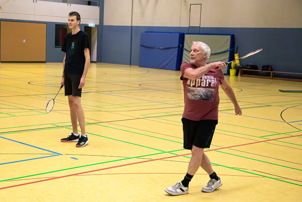 Zwei Männer spielen Badminton in einer Sporthalle. Der ältere Mann mit grauen Haaren trägt ein rotes T-Shirt und schwarze Shorts, während er einen Badmintonschläger schwingt. Der jüngere Mann steht in schwarzer Kleidung mit einem Schläger in der Hand und schaut aufmerksam zu. Der Boden der Halle ist gelb mit bunten Markierungen für das Badmintonspiel.