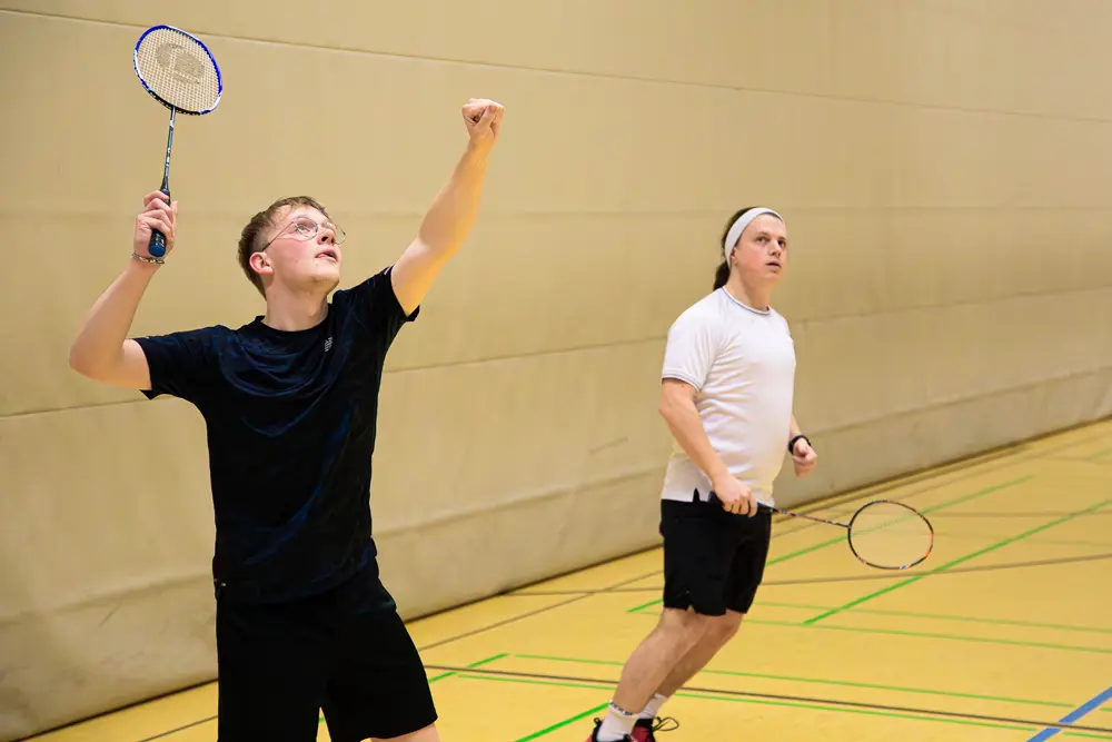 Zwei Badmintonspieler in einer Sporthalle, einer hebt seinen Arm mit dem Schläger, während der andere sich in Bewegung setzt.