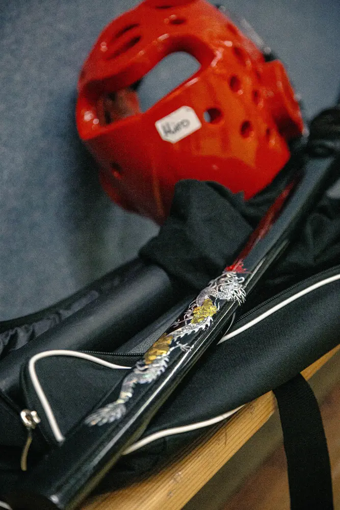 Ein roter Schutzhelm, eine schwarze Kummooyeh-Waffe mit Drachenverzierung und eine Sporttasche sind auf einer Holzoberfläche angeordnet, was auf die koreanische Kampfkunst hinweist.