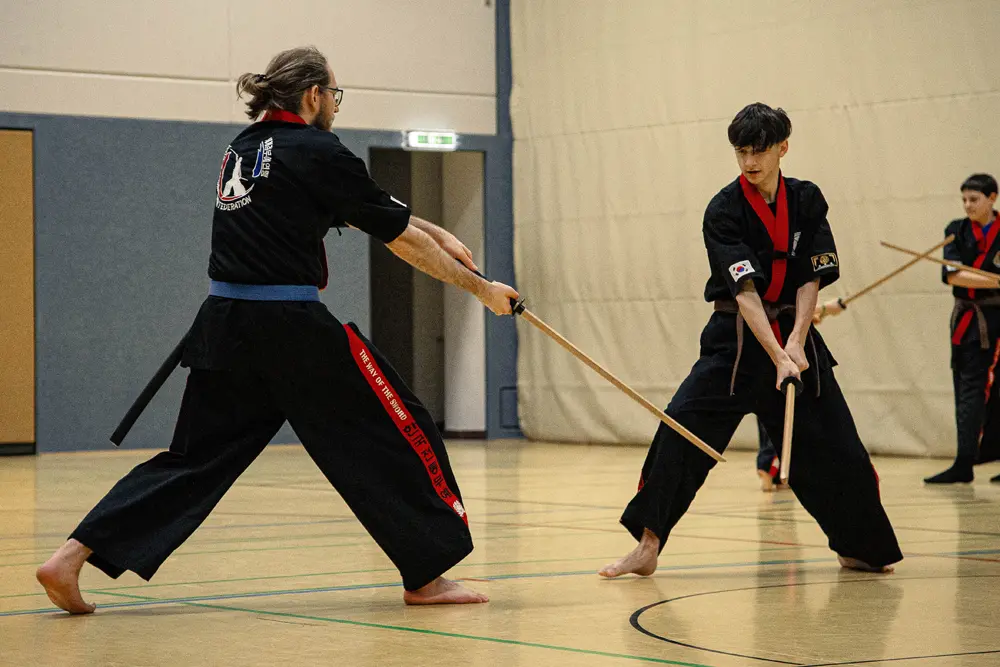 Zwei Kämpfer in schwarzer Kampfkunstbekleidung üben Kummooyeh in einer Halle. Der Kämpfer links hat einen blauen Gürtel und führt einen Stockkampf mit dem Kämpfer rechts, der einen roten Gürtel trägt. Im Hintergrund sind weitere Teilnehmer zu sehen, die ebenfalls mit Stöcken trainieren.