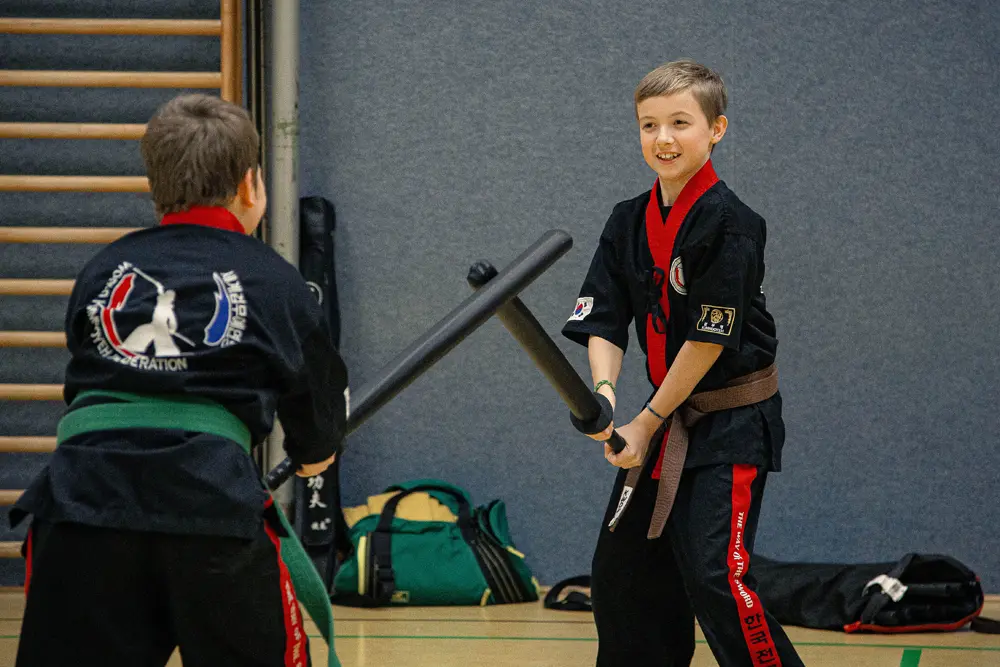 Zwei Kinder in Kampfkunstanzügen üben Kummooyeh mit Trainingsstäben in einer Sporthalle. Einer der Jungen hat einen grünen Gürtel und der andere einen braunen Gürtel.