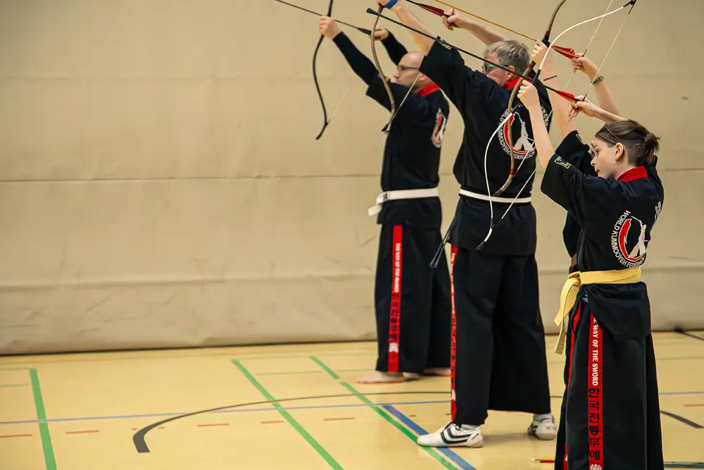 Drei Praktizierende der koreanischen Kampfkunst Kummooyeh stehen in einer Sporthalle und halten Bögen in der Zielposition. Sie tragen schwarze Trainingsanzüge mit farbigen Akzenten und Gürteln.