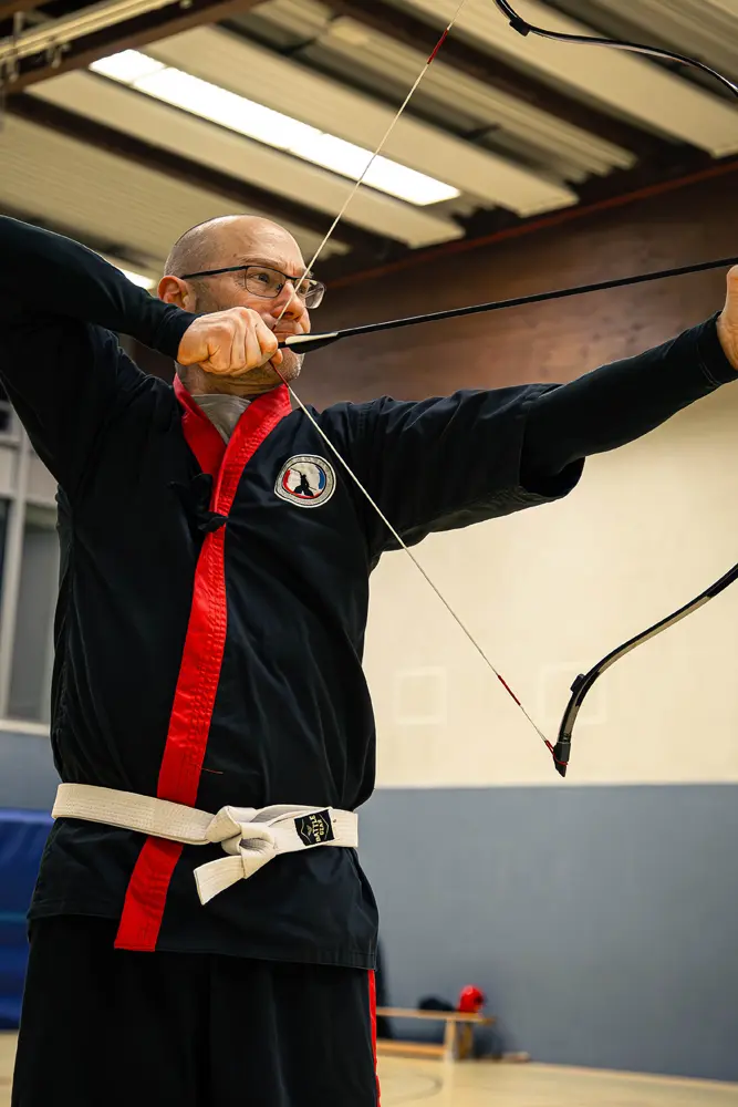 Ein Mann in einer schwarzen Kampfkunstuniform mit rotem Kragen und einem weißen Gürtel übt Kummooyeh, während er einen Bogen spannt.