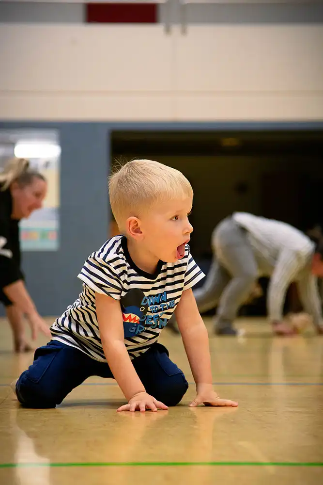 Ein kleiner Junge in gestreiftem T-Shirt und blauen Hosen krabbelt auf dem Boden einer Turnhalle, während andere Erwachsene im Hintergrund ebenfalls aktiv sind.
