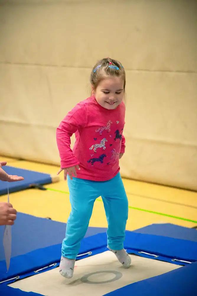 Ein Mädchen springt auf einem Trampolin während des Eltern-Kind-Turnens. Sie trägt ein pinkes Oberteil und blaue Hosen.