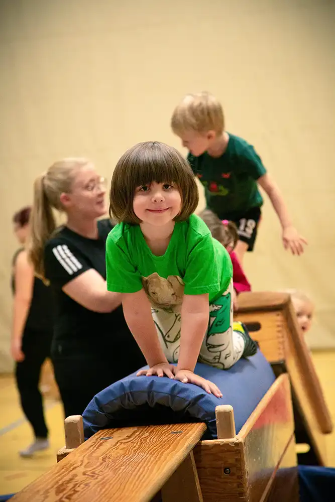 Ein Kind in einem grünen T-Shirt übt auf einer Turnbank, während andere Kinder und eine Trainerin im Hintergrund aktiv sind. Das Bild zeigt eine lebhafte Umgebung des Eltern-Kind-Turnens und Kinderturnens.