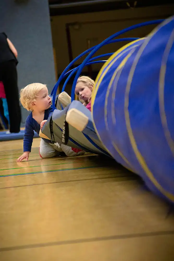 Zwei kleine Kinder spielen auf einer Turnmatte, während sie durch einen blauen Tunnel krabbeln. Das Bild zeigt eine aktive Umgebung für Eltern-Kind-Turnen und Kinderturnen.