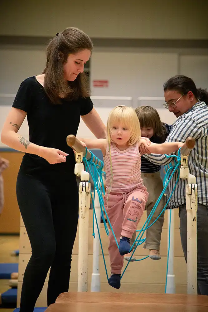 Ein Kind, das an einem Turngerät mit blauen Seilen spielt, während es von einer Erwachsenen unterstützt wird.