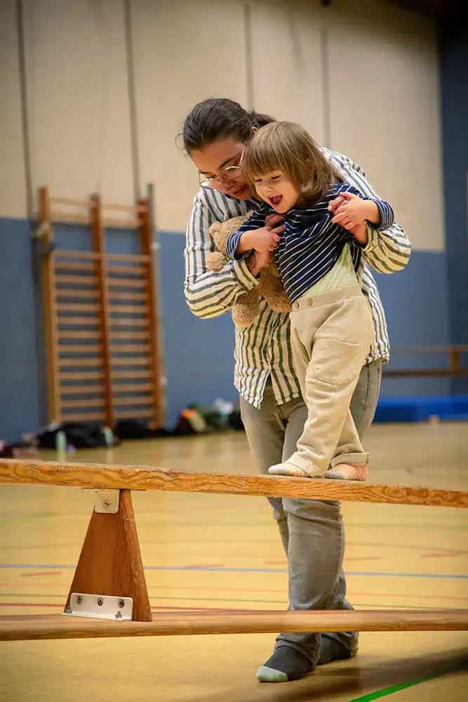 Eine Erwachsene hilft einem Kind, auf einem Turnbalken zu balancieren, während sie Spaß beim Eltern-Kind-Turnen haben.