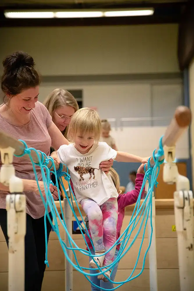 Ein Mädchen mit blonden Haaren balanciert auf einem Turngerät, während eine Frau sie unterstützt. Im Hintergrund sind weitere Frauen und Kinder zu sehen, die am Eltern-Kind-Turnen teilnehmen.