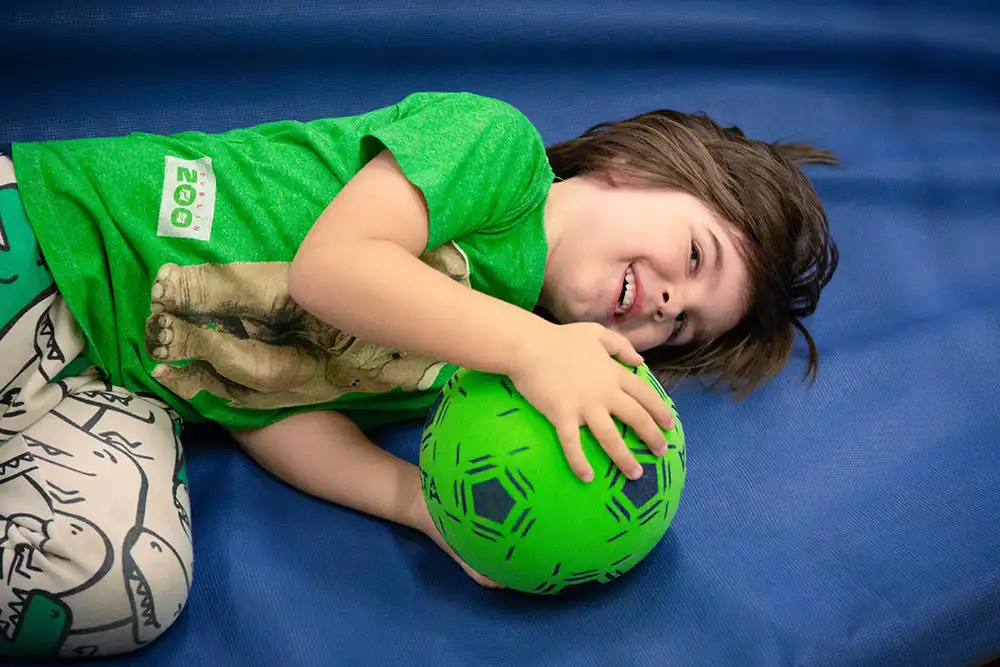 Ein lächelndes Kind liegt auf einer Matte und umarmt einen grünen Ball.