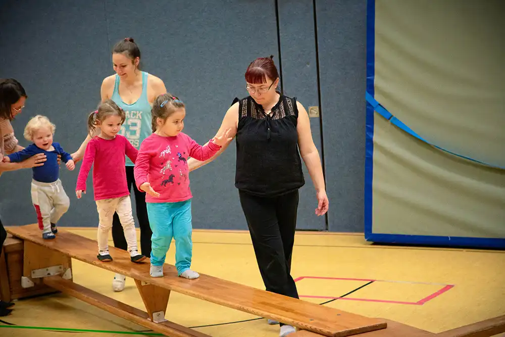 Kinder und Erwachsene beim Eltern-Kind-Turnen auf einem Balken; zwei Mädchen gehen auf dem Balken, während Erwachsene sie unterstützen.