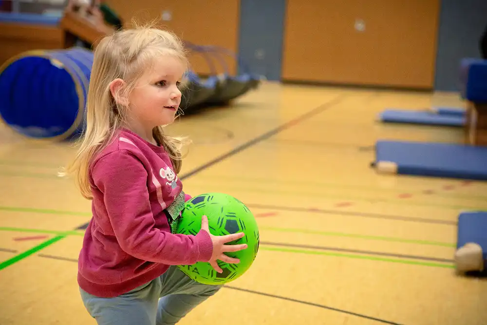 Ein kleines Mädchen mit langen blonden Haaren hält einen grünen Ball in der Hand. Die Atmosphäre ist freundlich und spielerisch, ideal für das Eltern-Kind-Turnen.