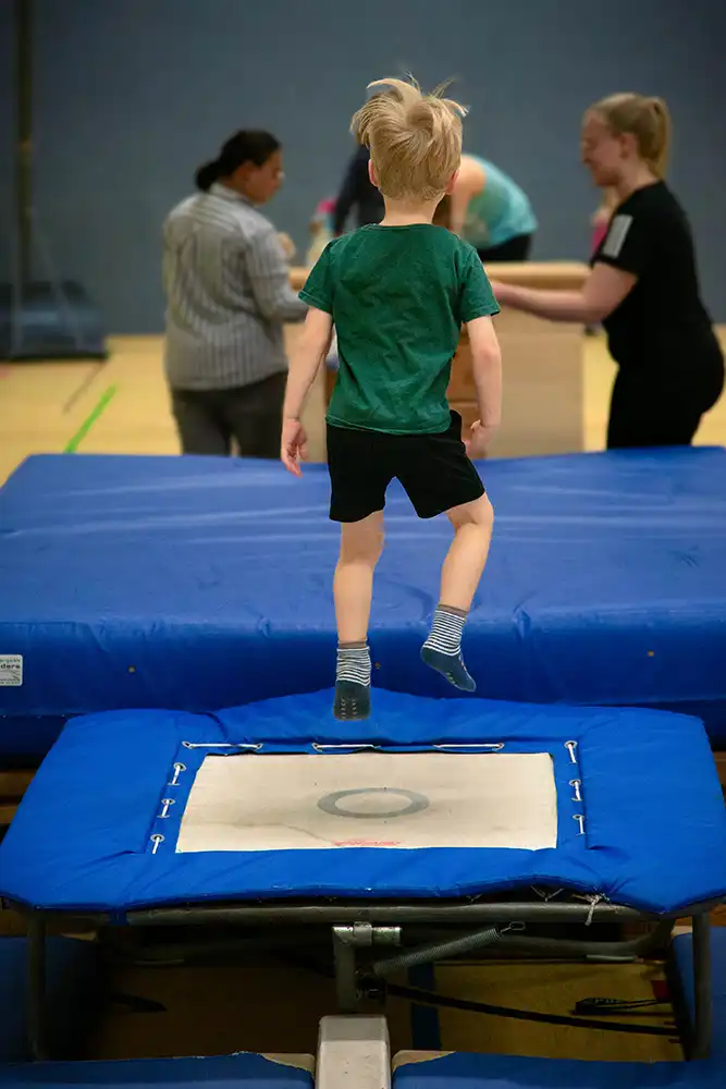 Ein Kind springt auf ein Trampolin während einer Eltern-Kind-Turnstunde, umgeben von anderen Teilnehmern in einer Turnhalle.