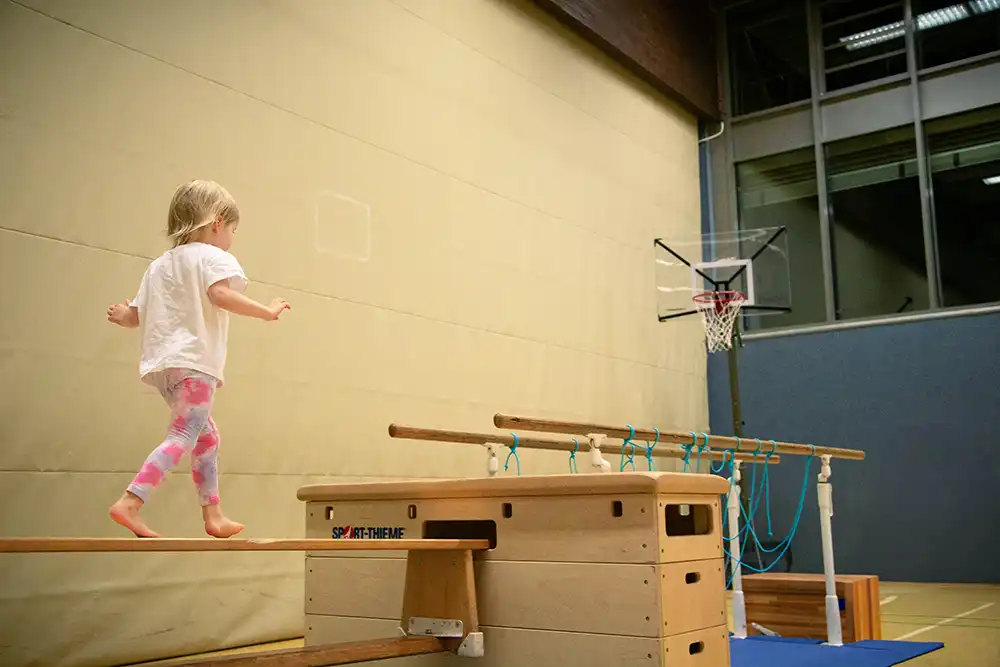 Ein kleines Kind balanciert barfuß auf einem Turnbalken in einer Turnhalle.